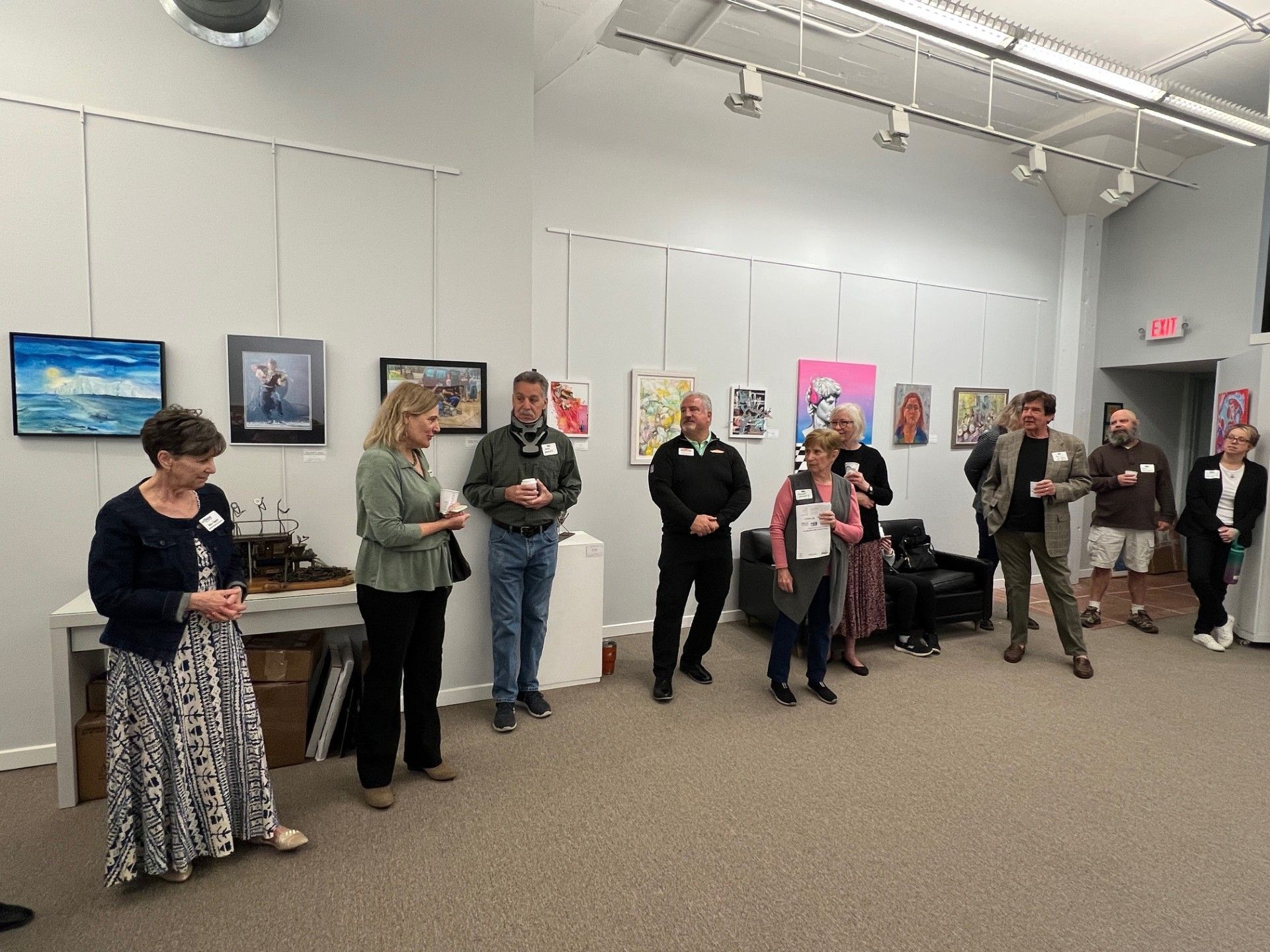 People standing in a gallery with artwork on the walls. Some are holding drinks, listening to a speaker.