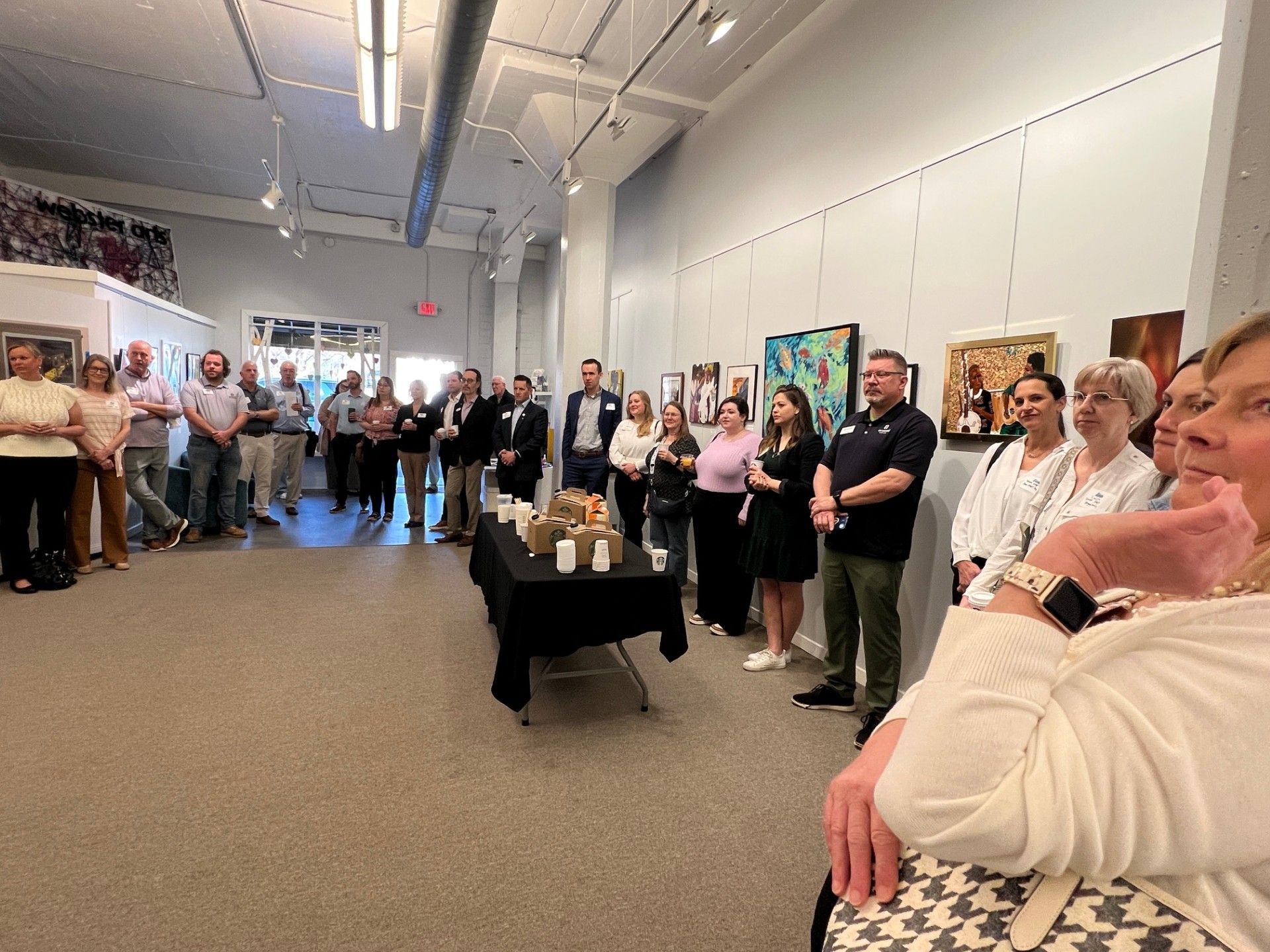 People gathered in an art gallery, viewing art on the walls. A speaker is at a small table, speaking to the group.