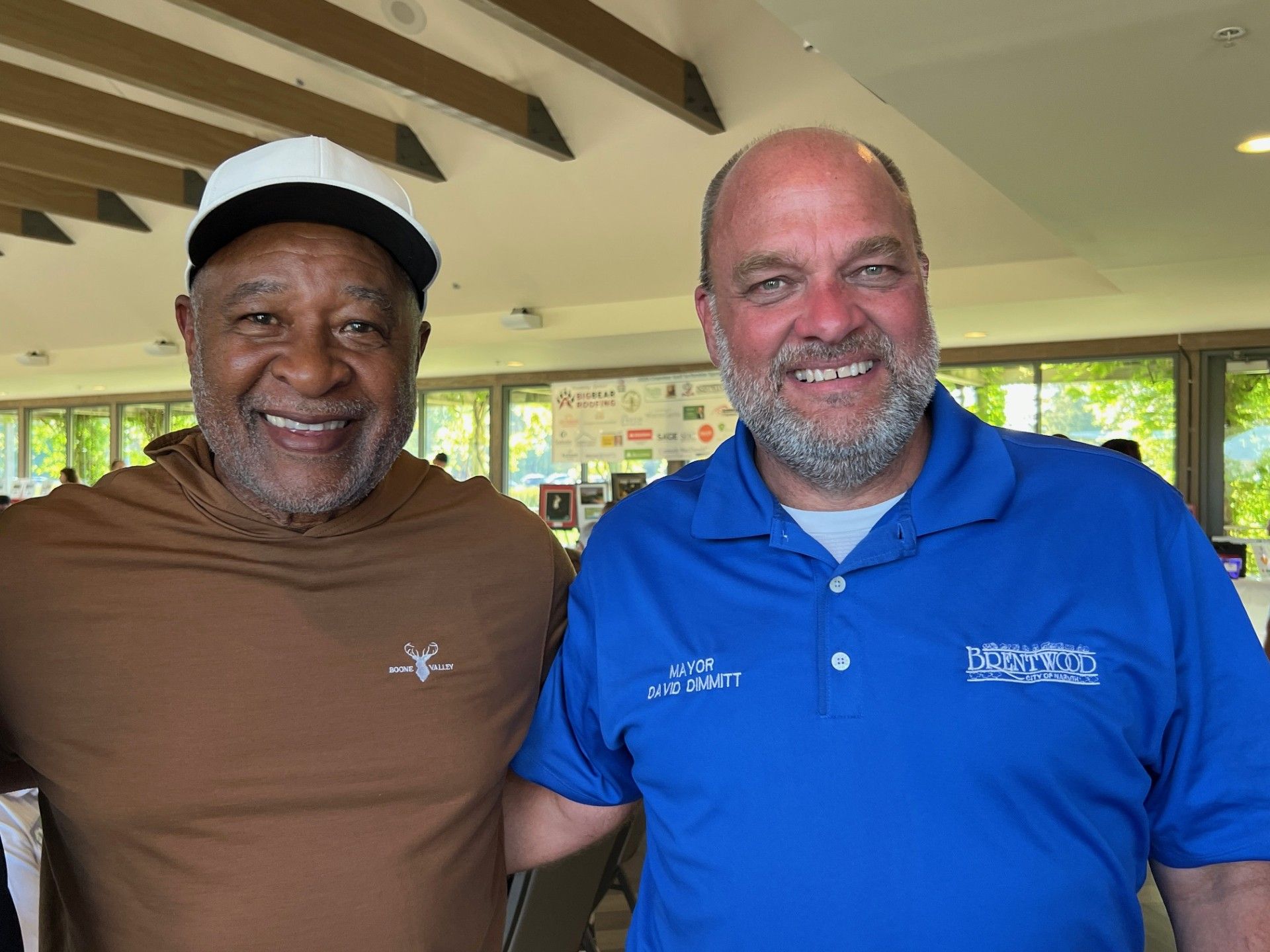 Two men smiling at the camera, posing outdoors. One wears a brown shirt and white hat, the other a blue polo.