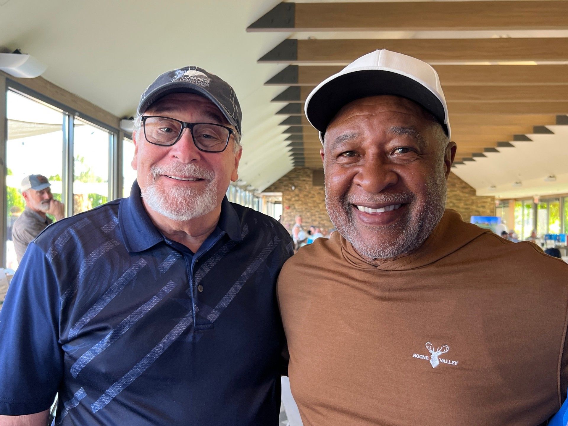 Two men smiling, one in a baseball cap, the other in a white-brimmed cap. They are outside.