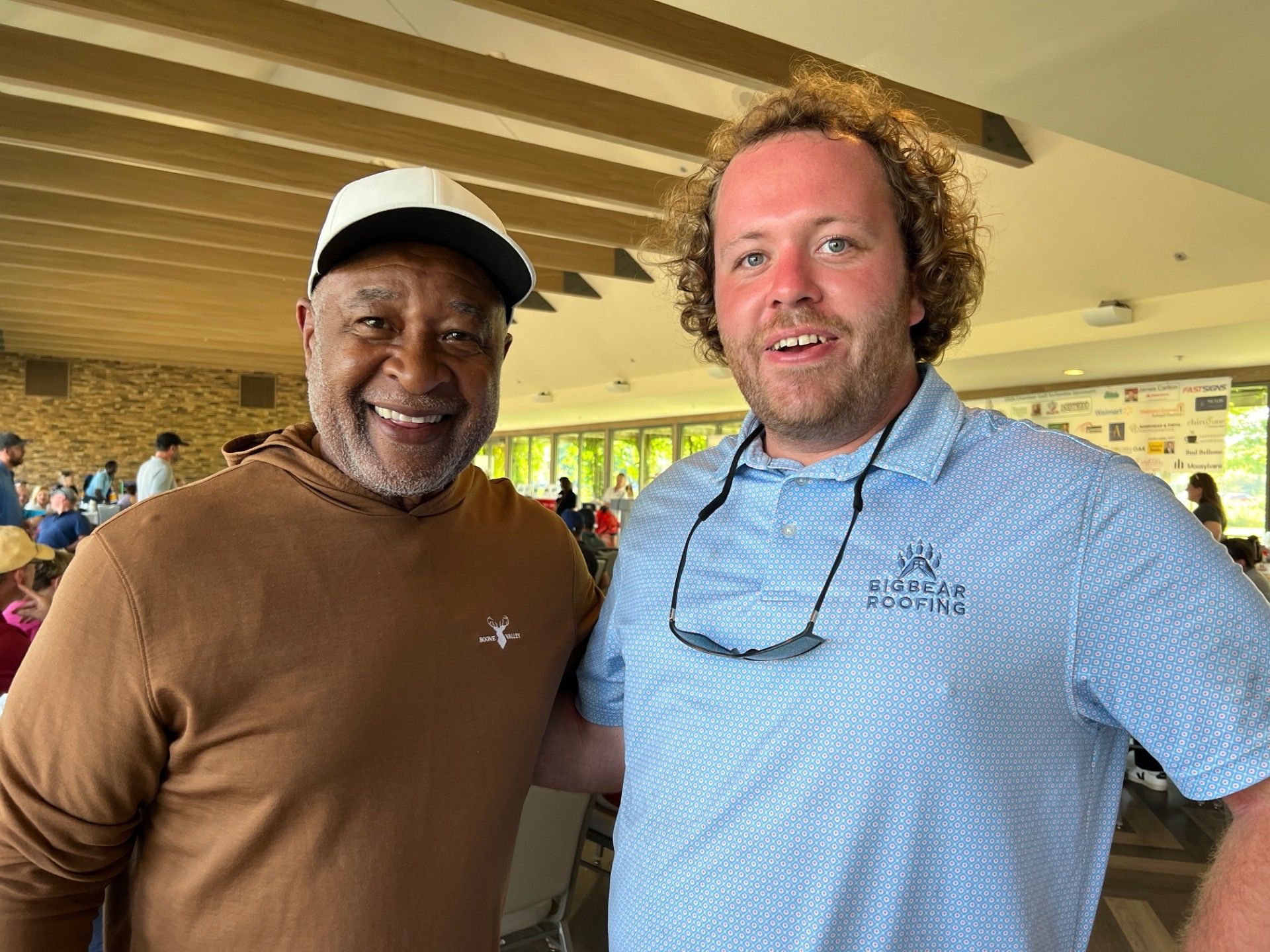 Two men smiling and posing for a photo, one wearing a brown hoodie and hat, the other a light blue polo.