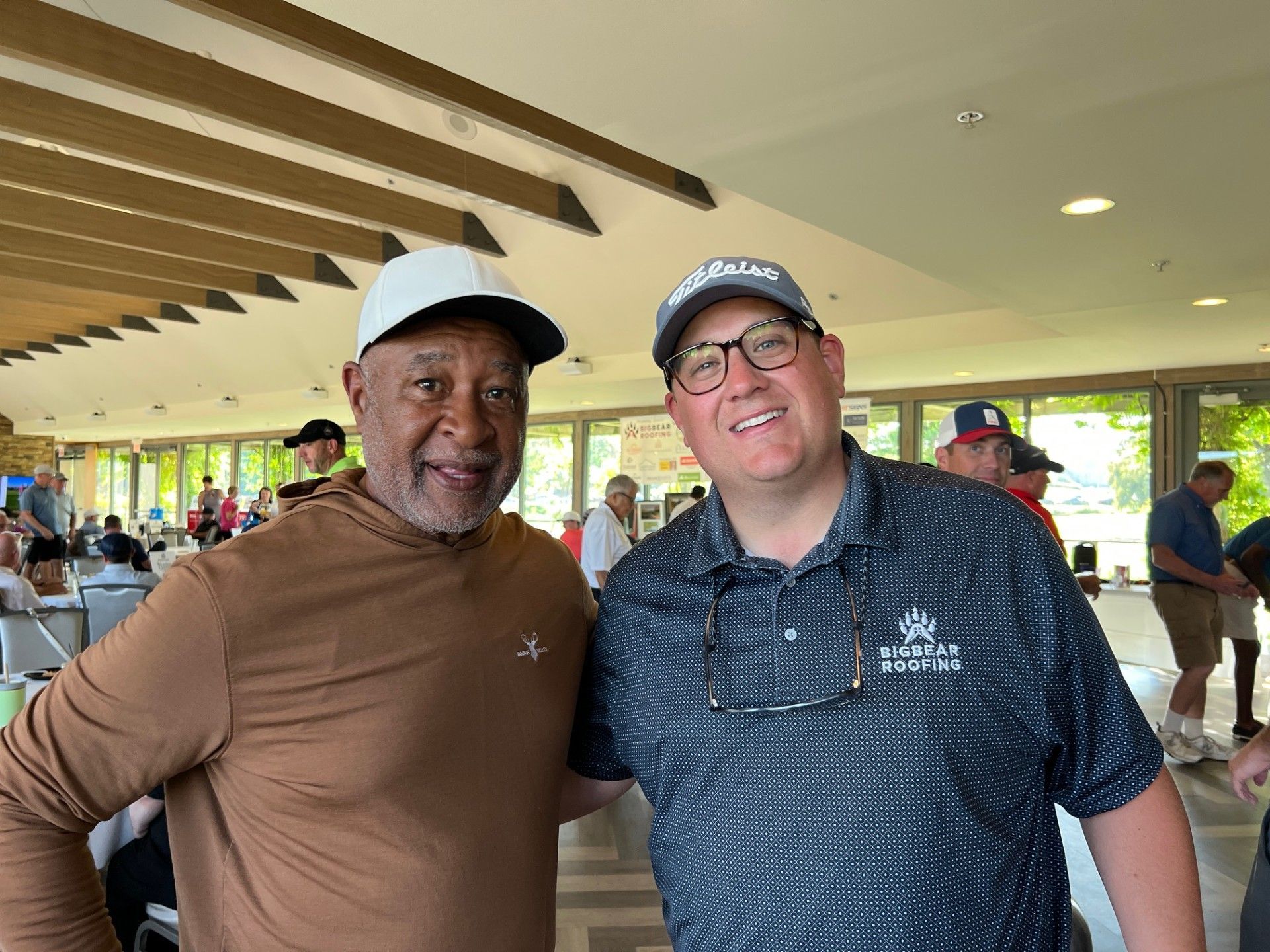 Two men smiling, posing together indoors. One in brown shirt and white hat, the other in a patterned blue shirt, Titleist hat, and glasses.