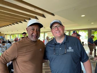 Two men smiling, posing together indoors. One in brown shirt and white hat, the other in a patterned blue shirt, Titleist hat, and glasses.