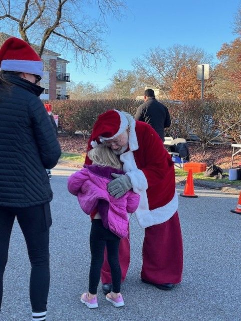 Santa Claus hugs a child wearing a pink jacket, watched by a person in a black jacket and a red hat, outdoors.