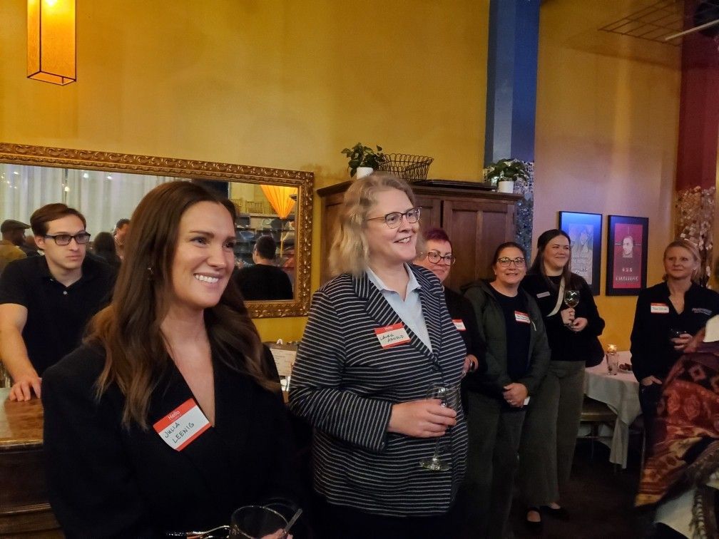 People at a gathering in a room with a mirror and bar. Some wear name tags and smile. Warm lighting.