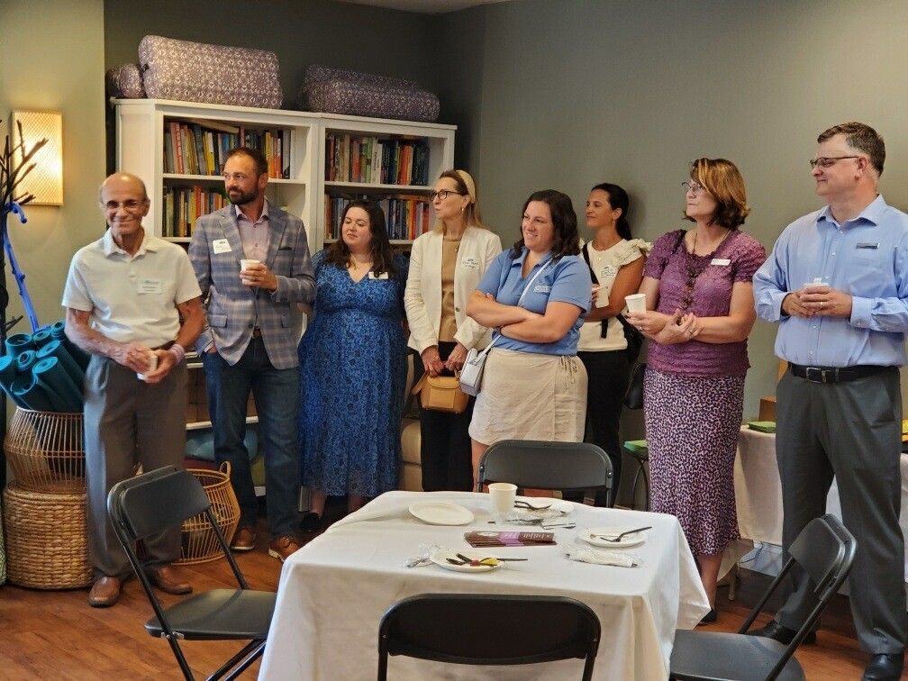 Group of people in a room standing near a table with food, looking toward the viewer. Bookshelves in the background.