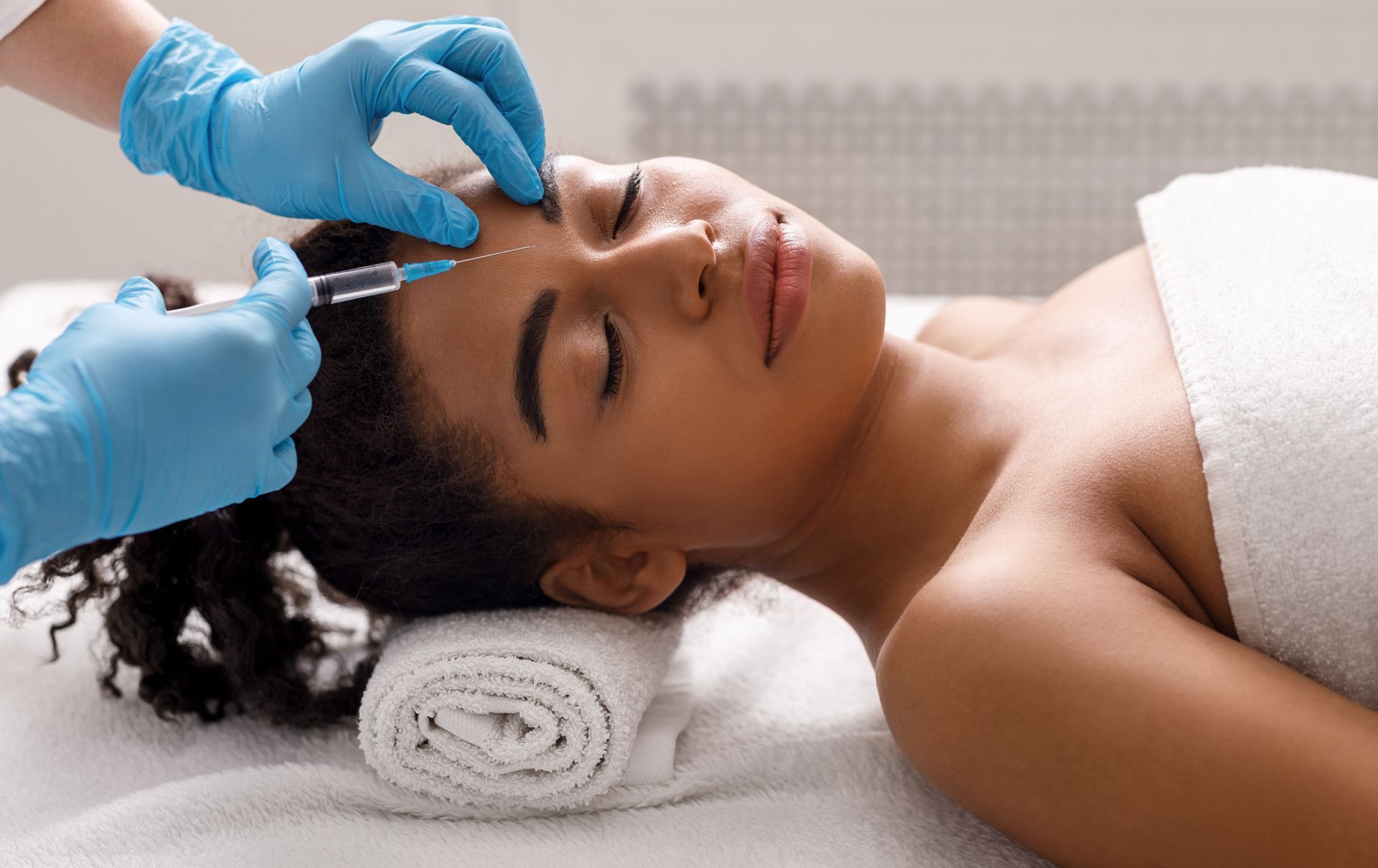 Person receiving a cosmetic injection in their forehead. Gloved hands holding syringe, on white spa bed.