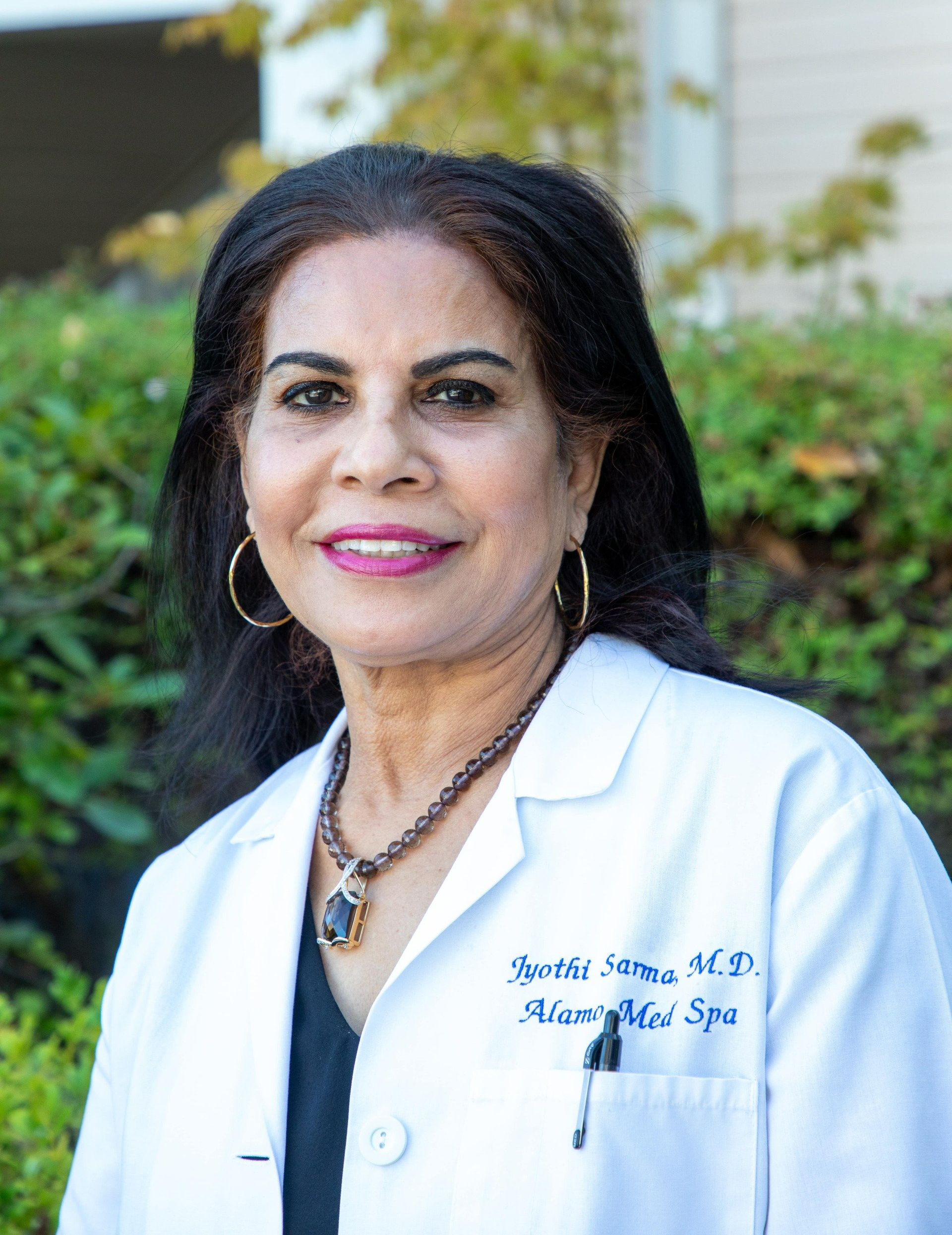 Dr. Sarma - Alamo, CA - Alamo MD Med Spa Woman in white coat, wearing necklace and earrings, smiles outdoors.