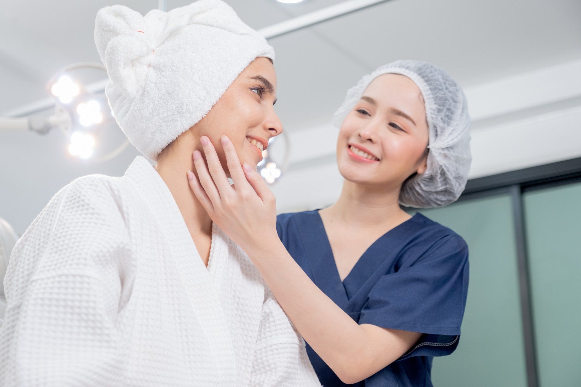 Woman in robe with towel on head, being examined by healthcare worker in surgical scrubs, smiling.