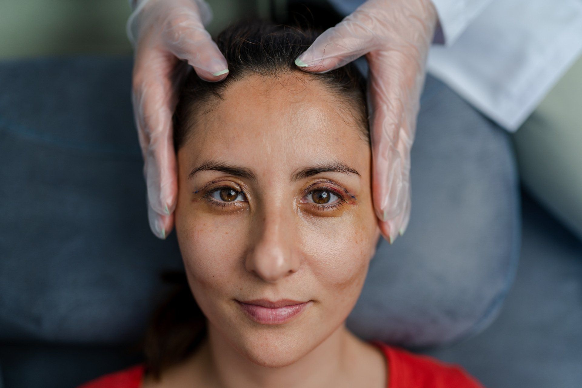 Person with hands on forehead. Medical gloves, red shirt, brown eyes.