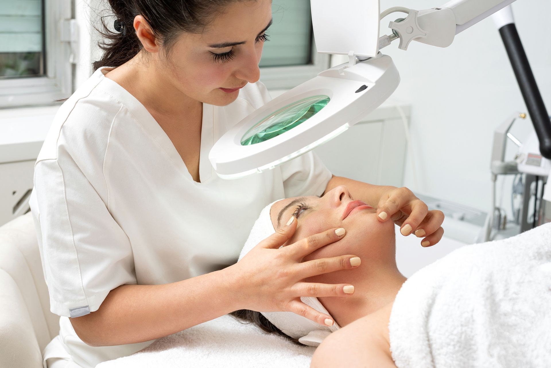 Woman receiving facial treatment from a person wearing a white uniform, under a magnifying lamp.