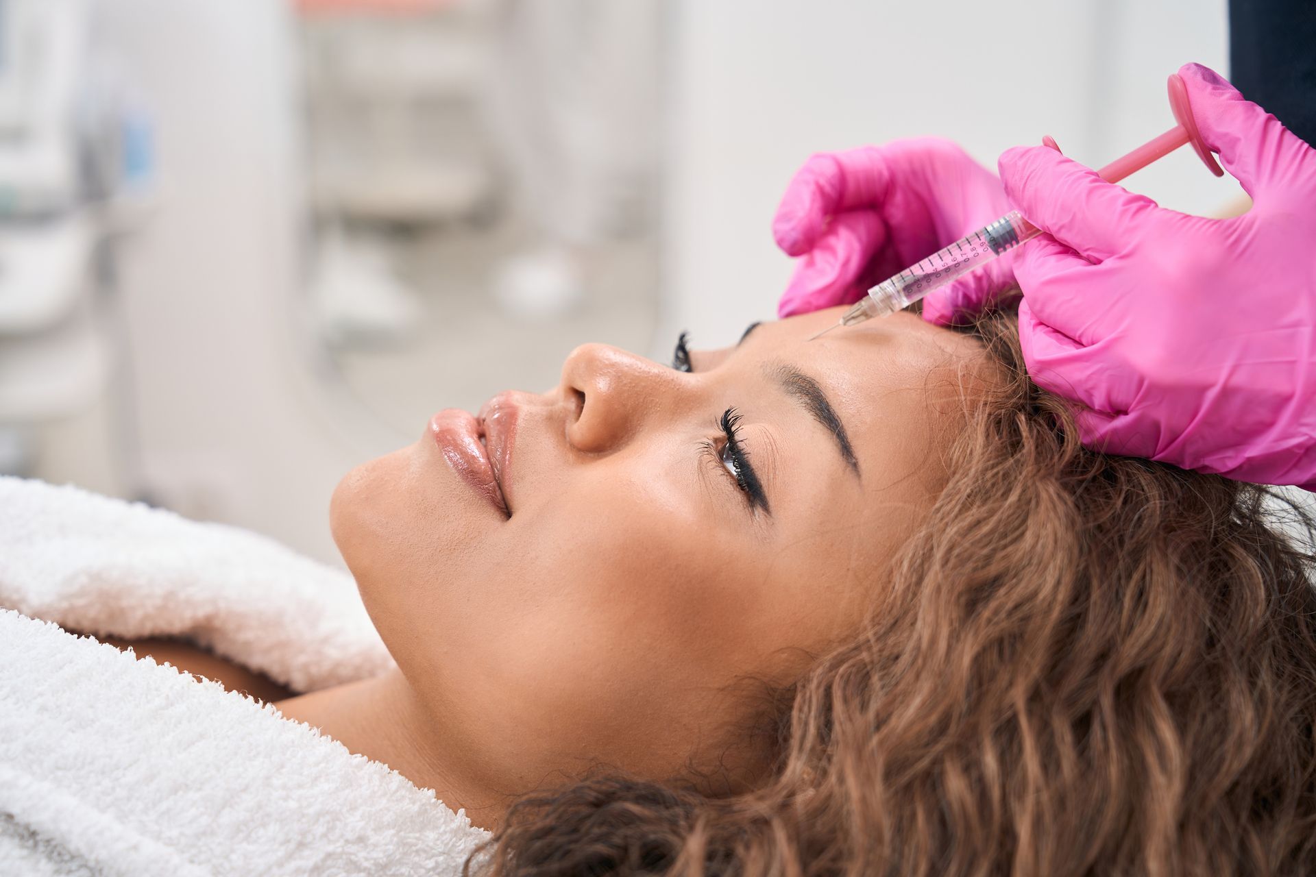Person receiving facial injections; pink-gloved hands holding a syringe, near the forehead.