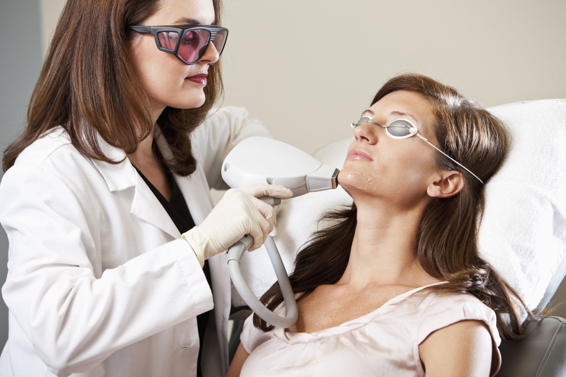 Woman receiving laser skin treatment on her neck from a professional. Both wear protective eyewear.