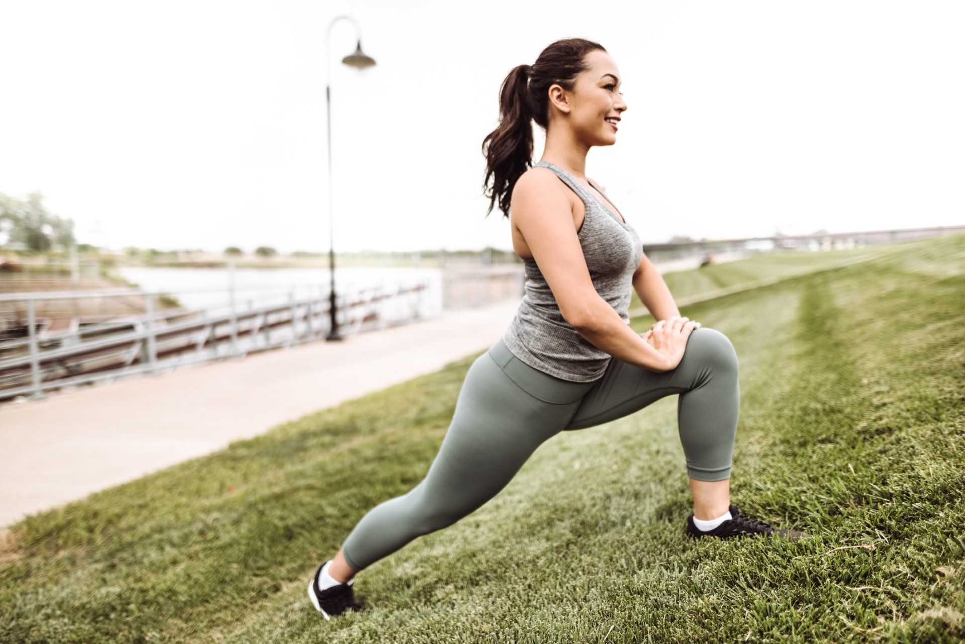 Woman in workout clothes lunging on a grassy hill, hands on knee, outdoors.