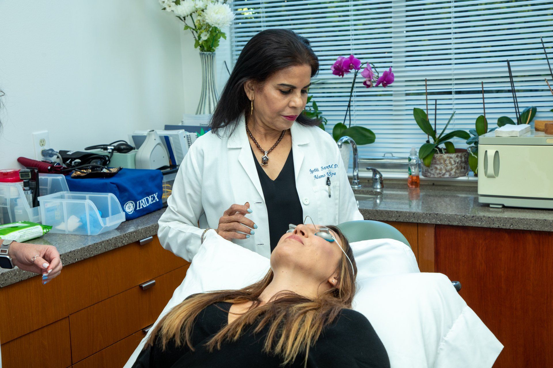Woman in lab coat preparing to treat a person lying down on an exam table. Flowers and equipment in the background.
