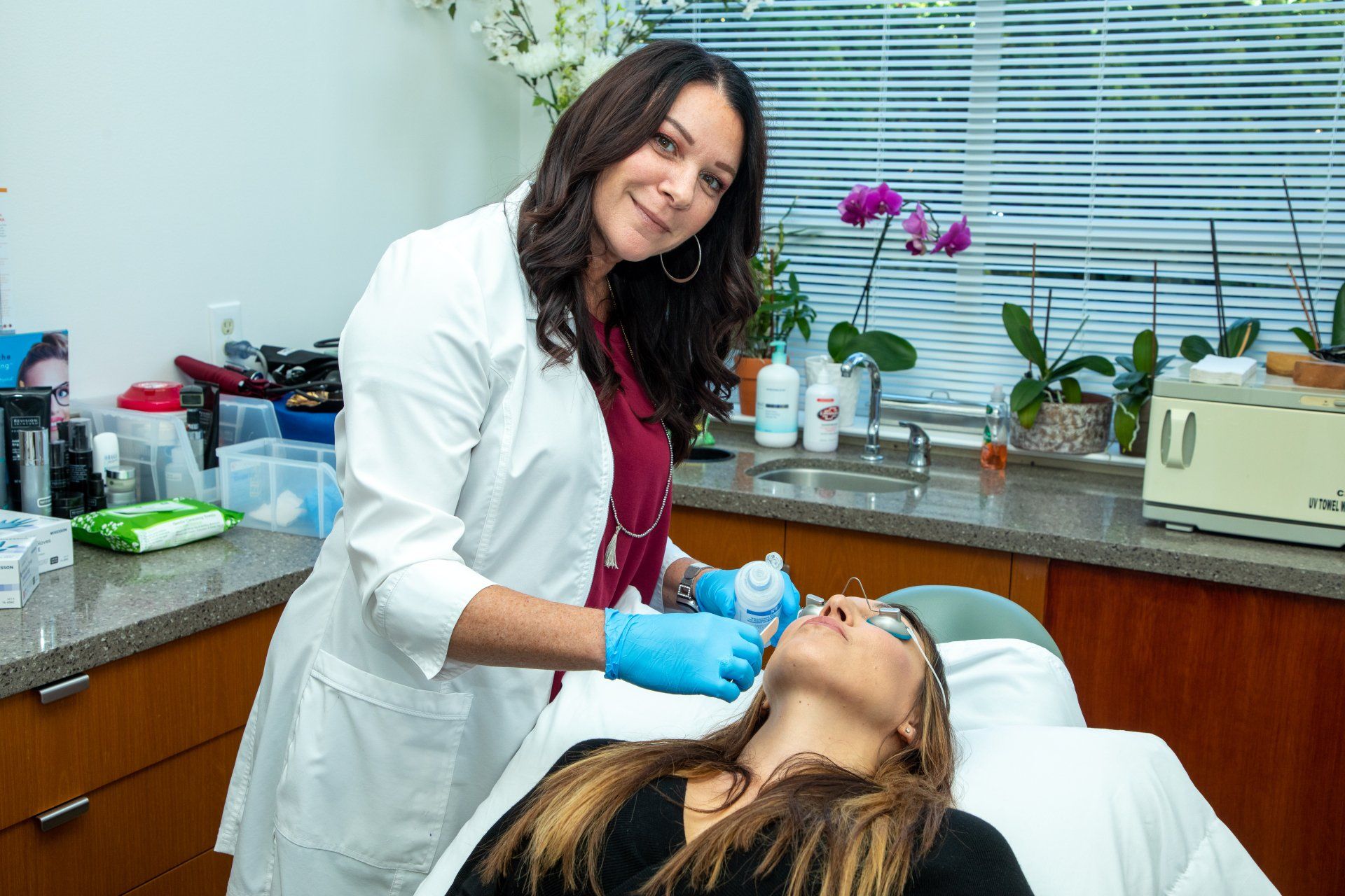 Woman in white coat administering cosmetic treatment to another woman's face.