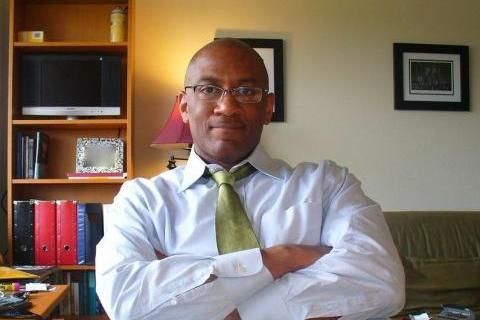 Man in white shirt and green tie sitting with arms crossed in an office, bookshelf behind him.