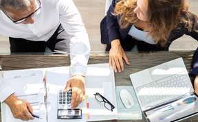 Two people leaning over a desk, reviewing documents with a calculator, glasses, and a notebook.