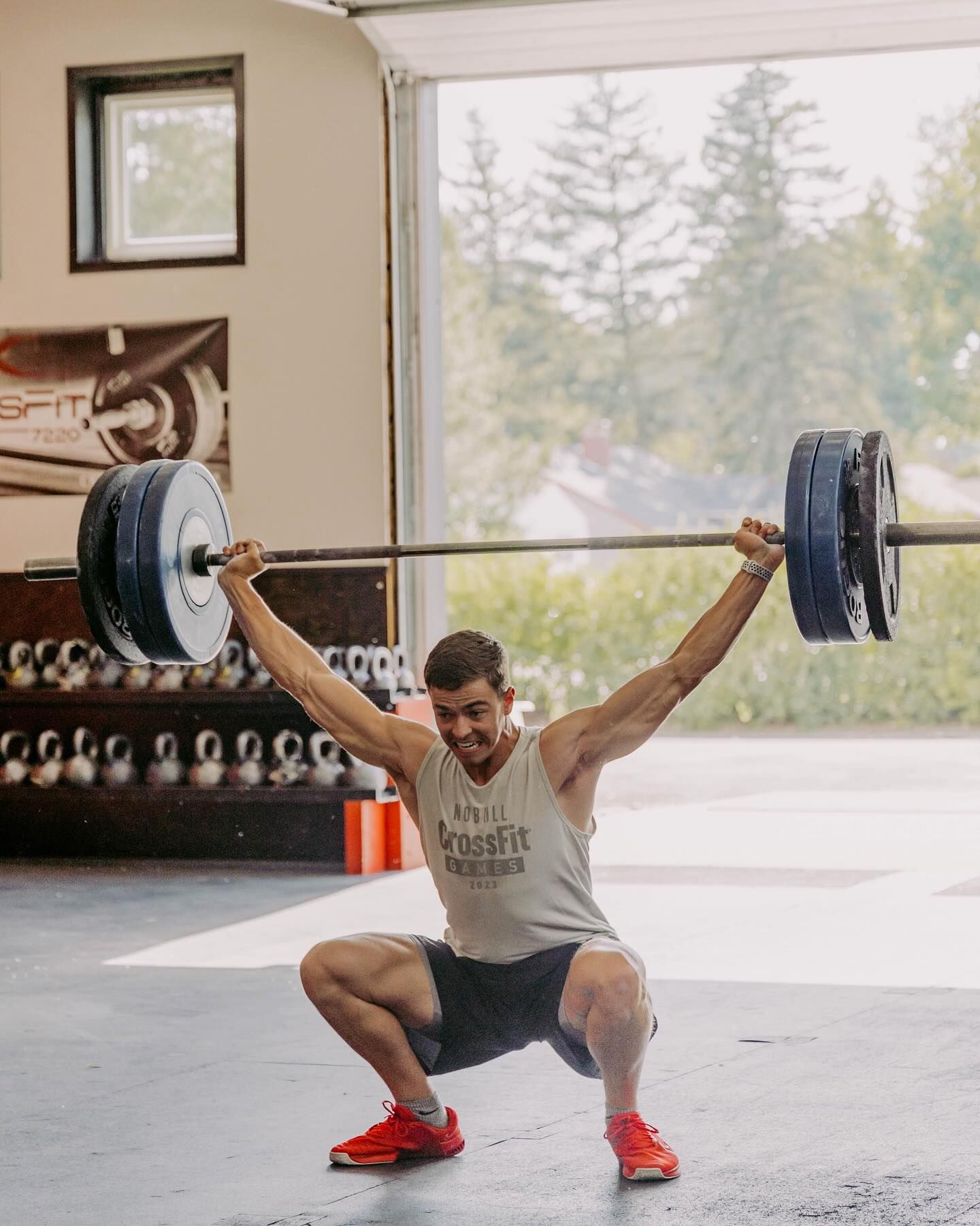 Man in a gym overhead squatting with a barbell, arms extended. Sunlight shines through the open garage door.