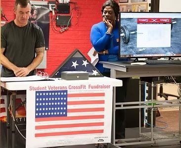 Man and woman at a Student Veterans CrossFit fundraiser, standing near an American flag and a presentation screen.