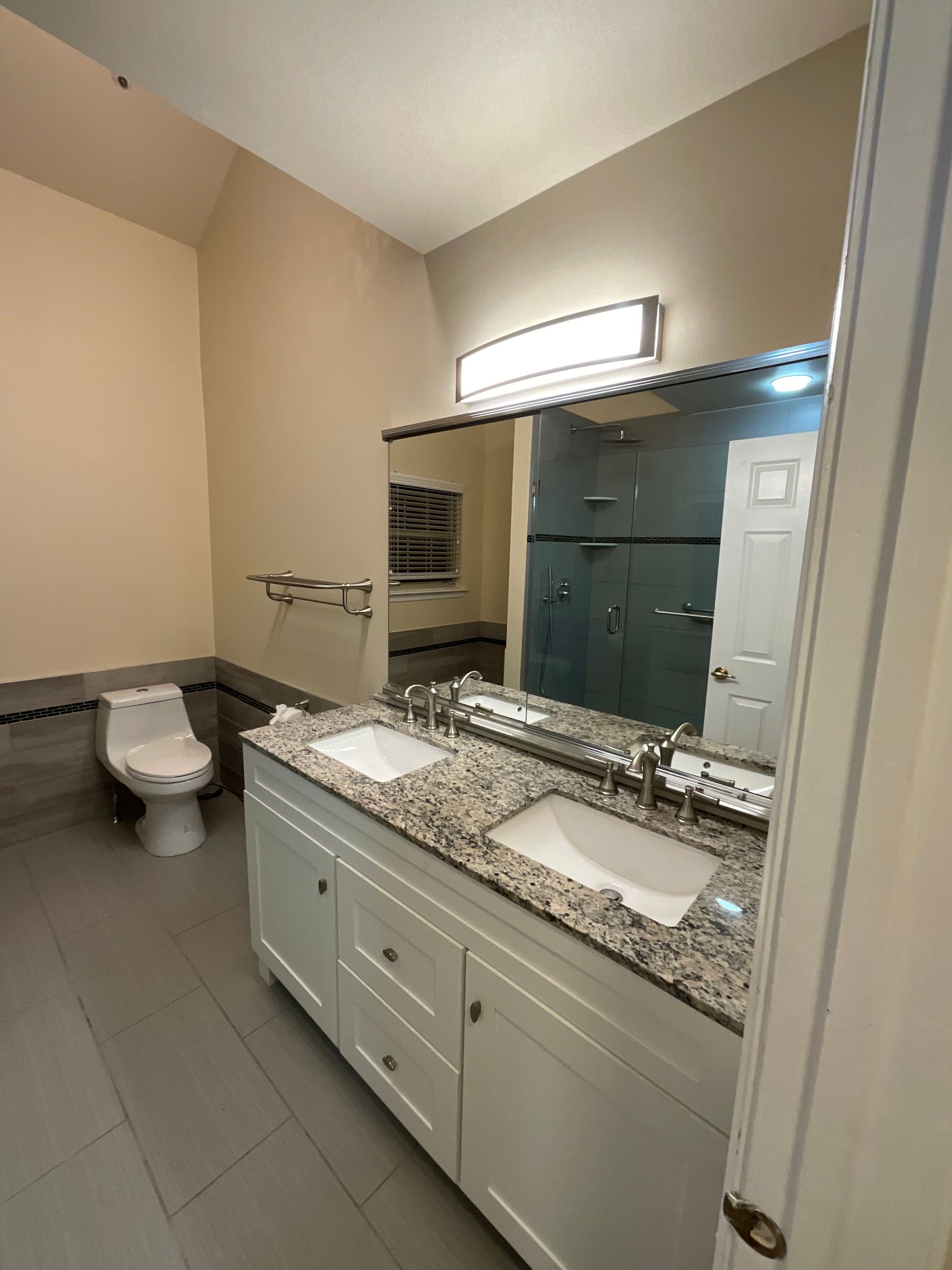 A modern bathroom featuring a white dual-sink vanity with a speckled stone countertop, a large mirror, and a toilet.