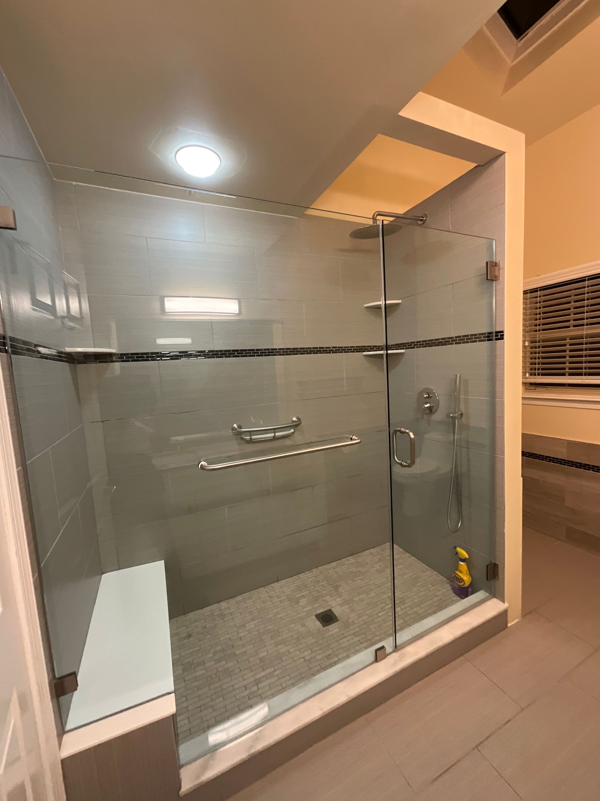 A modern walk-in shower with a glass door, a built-in white bench, a horizontal grab bar, and neutral gray tile walls.