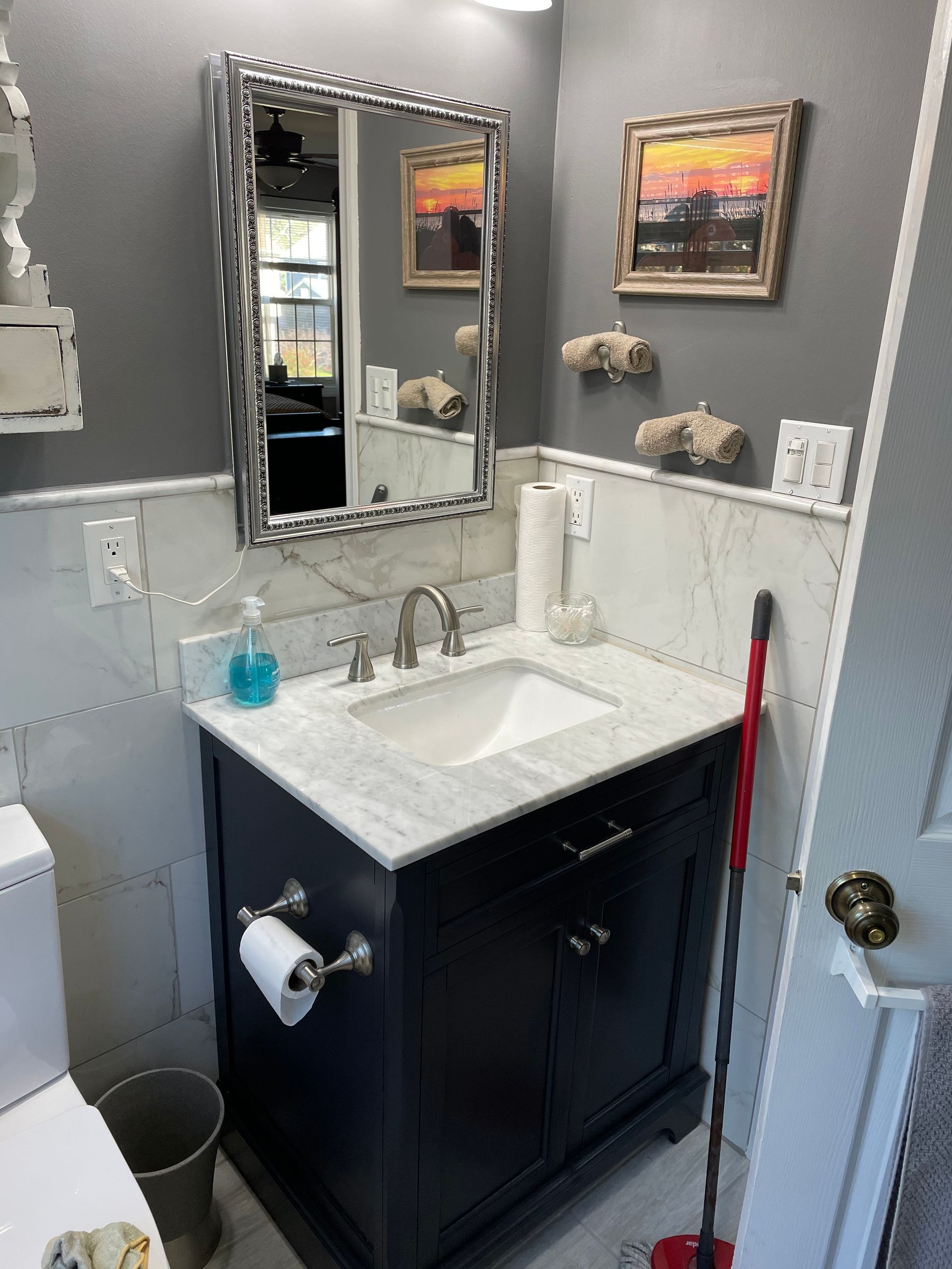 A bathroom vanity with a dark cabinet, white marble-style countertop, a mirror, and gray walls, with a plunger to the side.