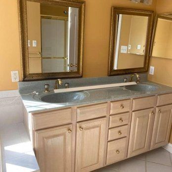 A double bathroom vanity with wood cabinets, light grey countertops, and two gold-framed mirrors against tan walls.