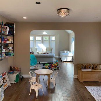 A playroom with a small table and chairs, connected by an archway to a bright, sunlit living area with sofa seating.