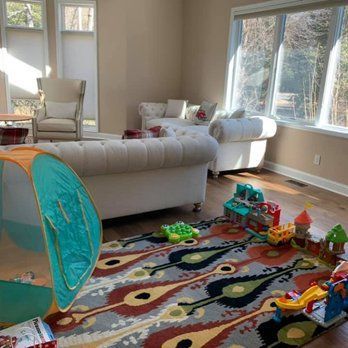 A bright living room features a white tufted sofa, armchair, and a colorful patterned rug with children's toys scattered.