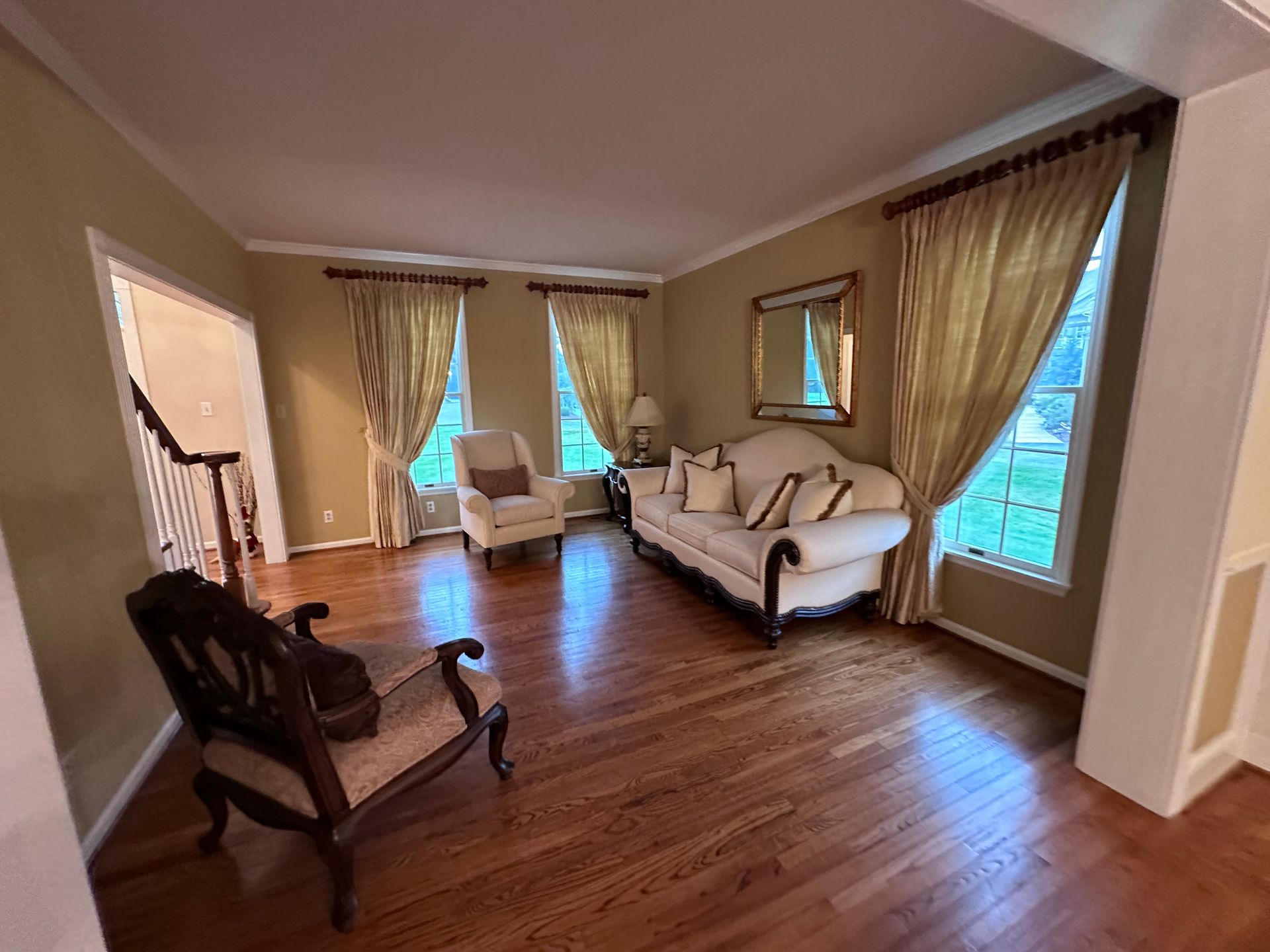 A living room with hardwood floors, a white sofa, two chairs, beige curtains, and a mirror on a tan wall.