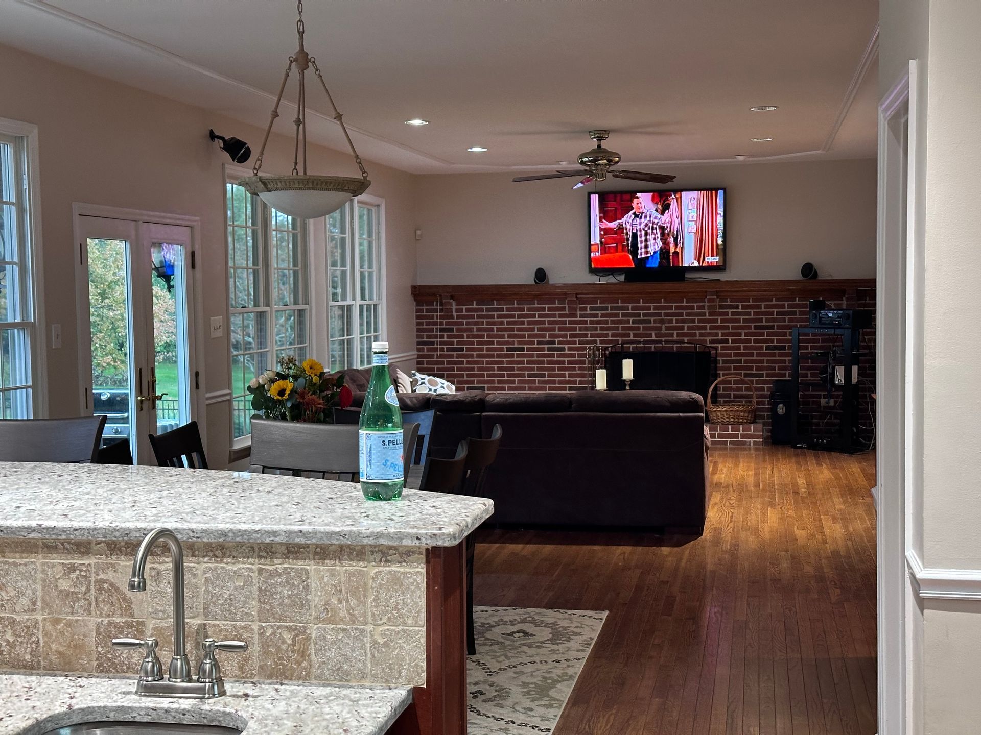 A kitchen island with a granite countertop and sink in the foreground, overlooking a living room with a brick fireplace.