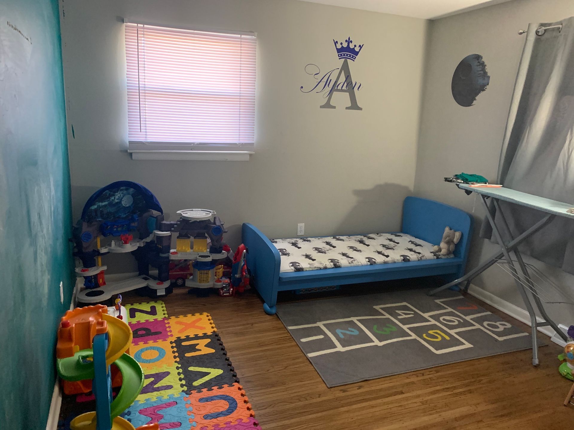 A child's bedroom features a blue toddler bed, a hopscotch rug, an alphabet play mat, and toys on a wood floor.
