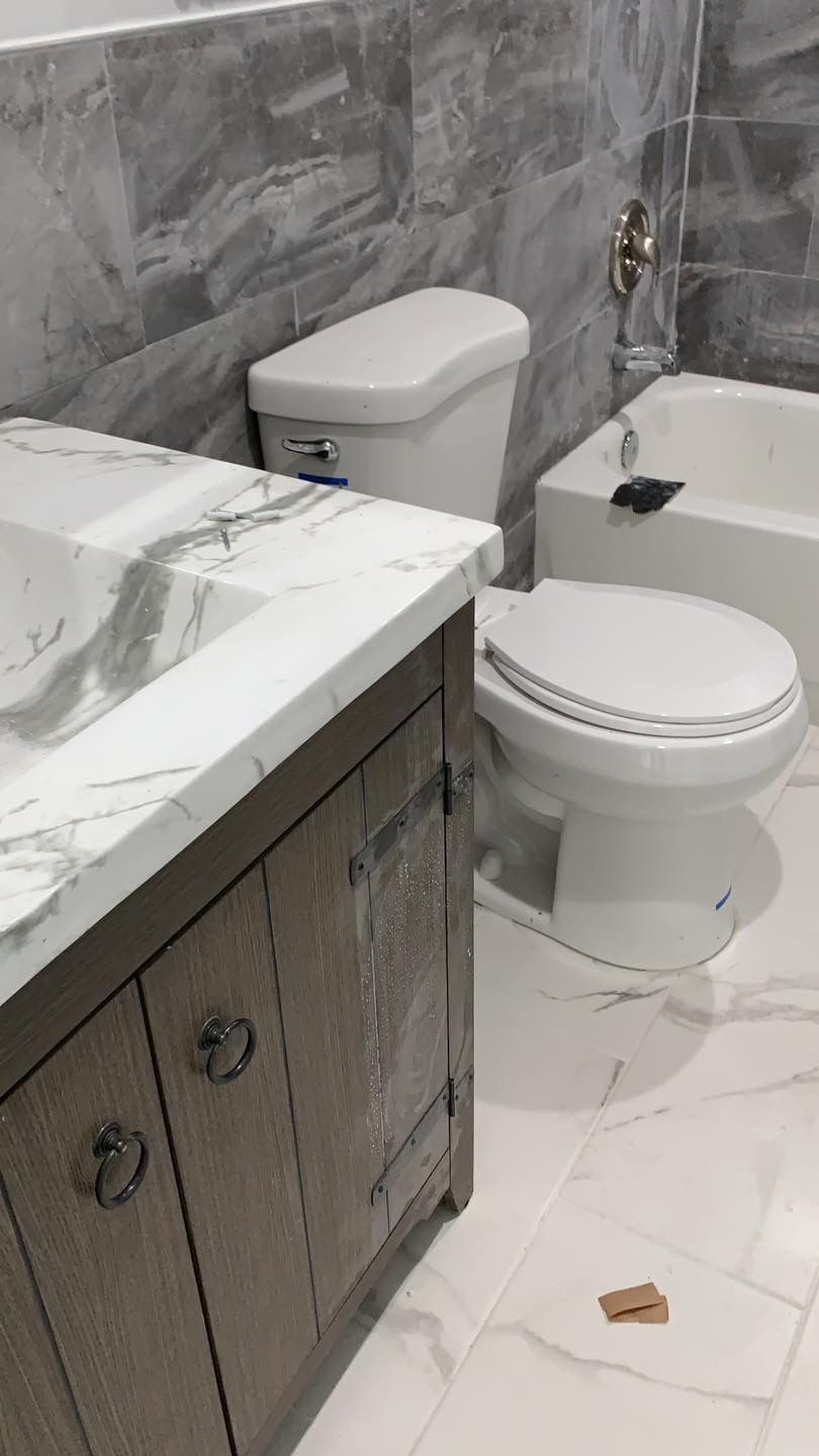 A modern bathroom with a wooden vanity, white marble countertop, white toilet, and gray tiled walls with a bathtub.