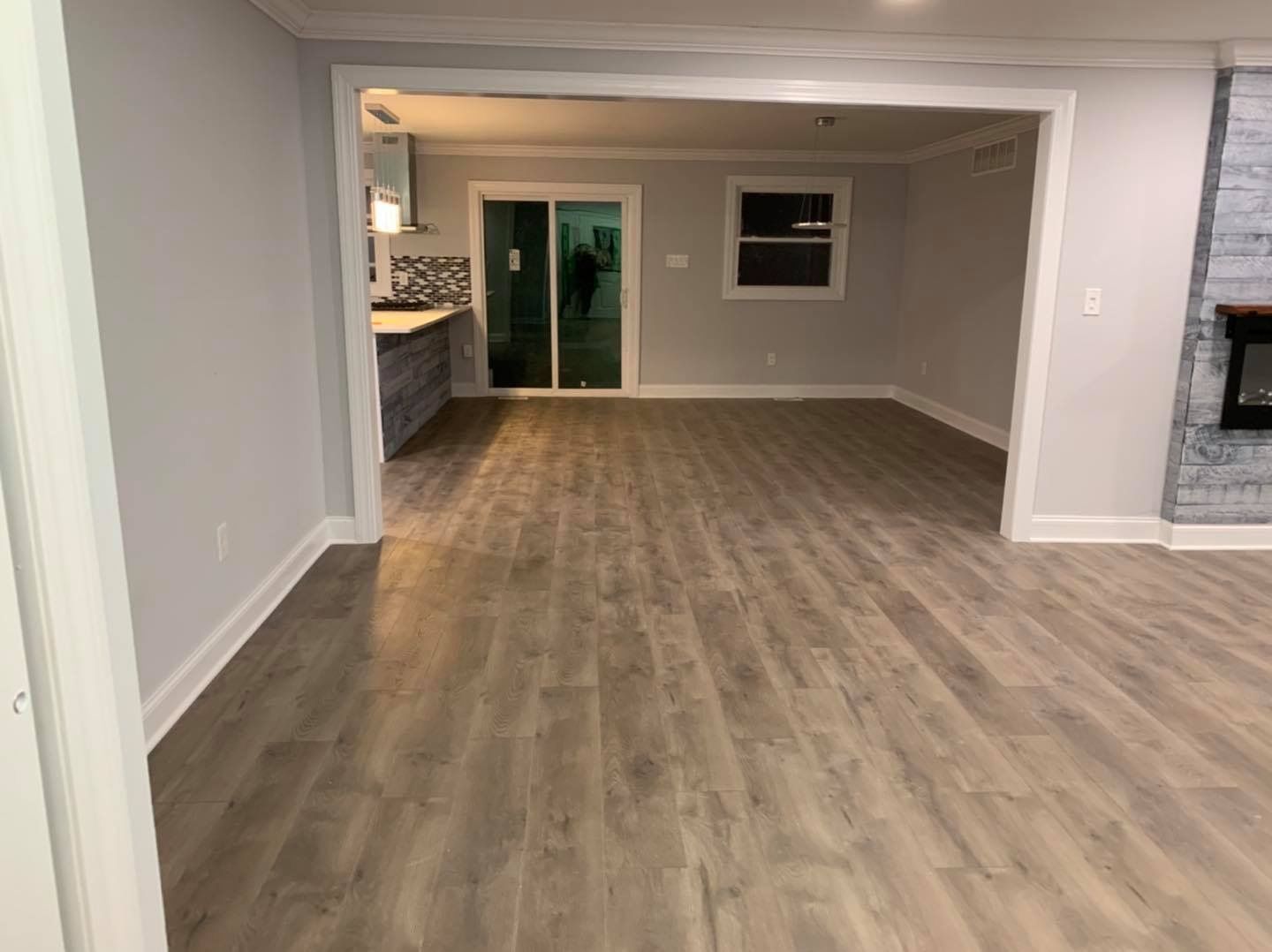 An open-concept living area with gray laminate flooring, light gray walls, and a view into a kitchen with a sliding door.