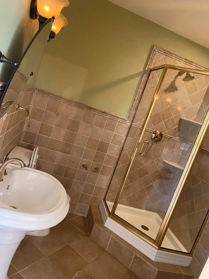 A bathroom with a pedestal sink, a corner shower with gold-toned framing, and tan-tiled walls against a light green wall.