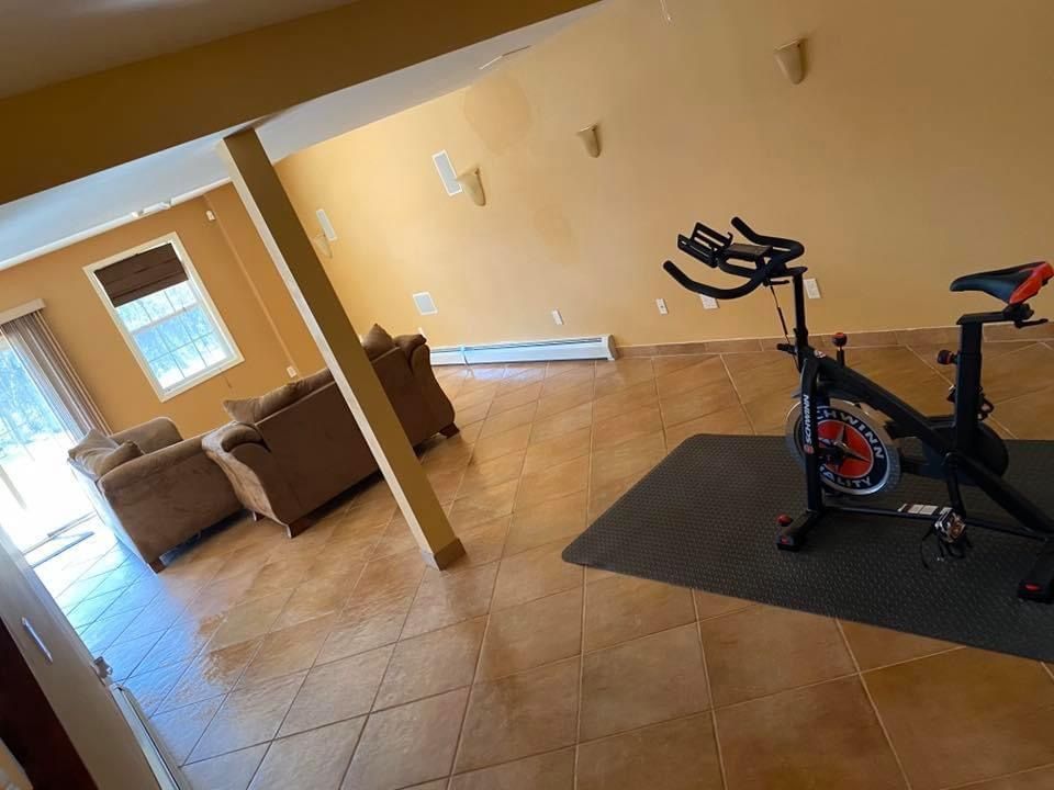 An exercise bike sits on a mat in a spacious, open-plan living room with tan tile floors and beige walls.