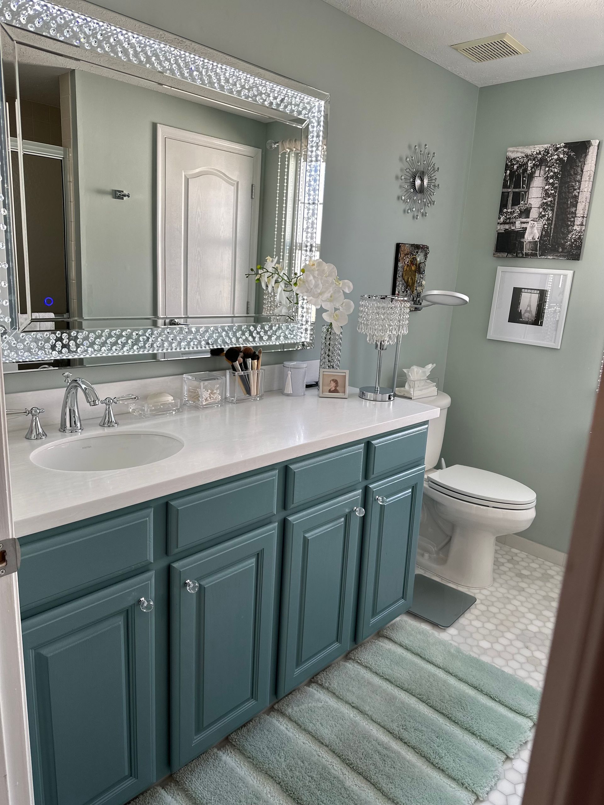 A modern bathroom with blue-grey vanity cabinets, a white countertop, large decorative mirror, and light green walls.