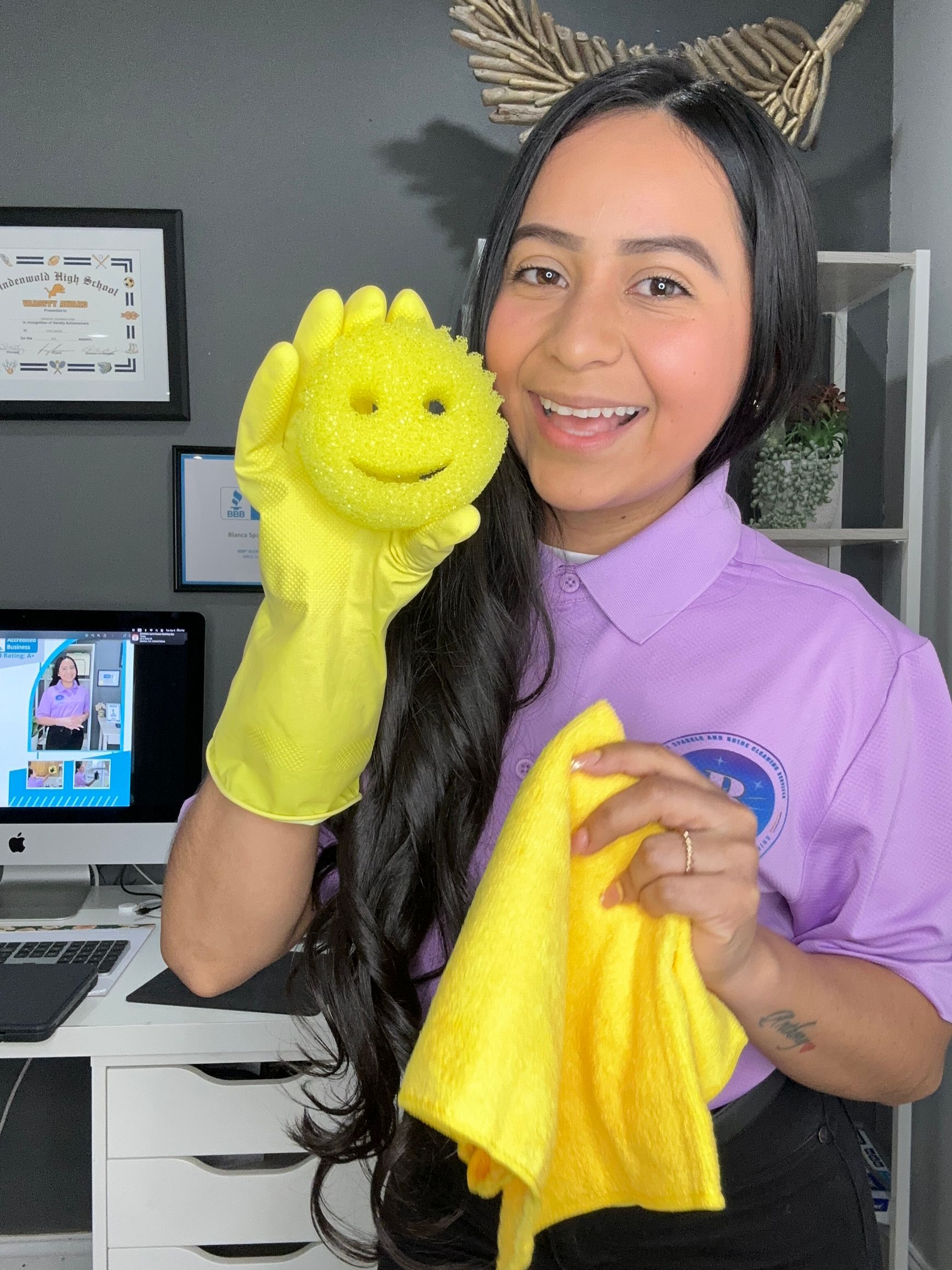 Smiling person in a purple shirt holding a yellow smiley-face sponge and a yellow cleaning cloth in a home office.