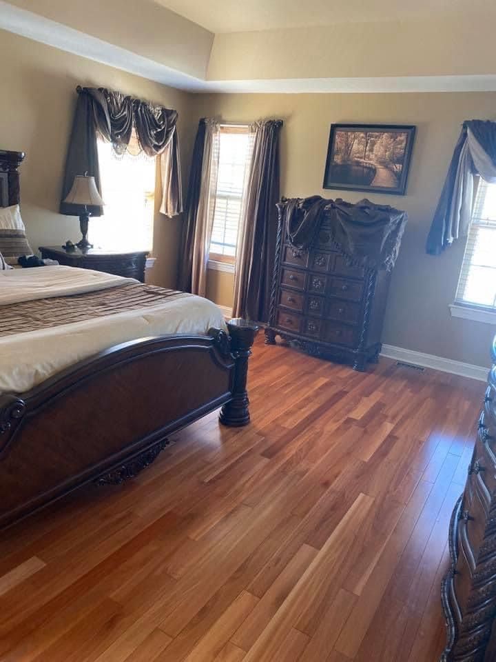 A bedroom with a dark wooden bed, a dresser, and two windows with layered curtains, all set on light wood floors.