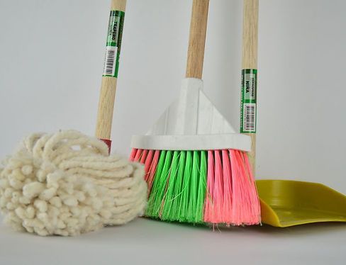 A cotton string mop, a broom with neon green and pink bristles, and a yellow dustpan standing on a white background.