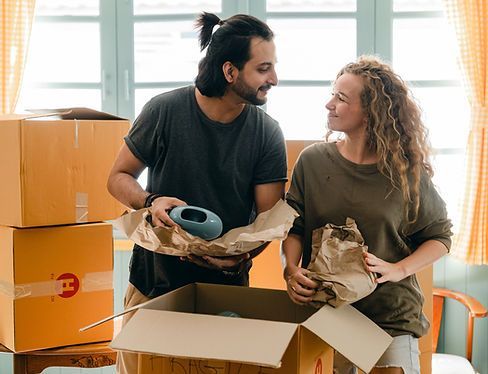 A couple in a room filled with cardboard boxes, wrapping an object in paper while looking at each other.