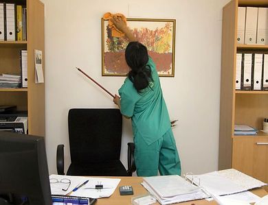A person in green scrubs uses a yellow cloth to clean a framed picture hanging on a white wall in an office.