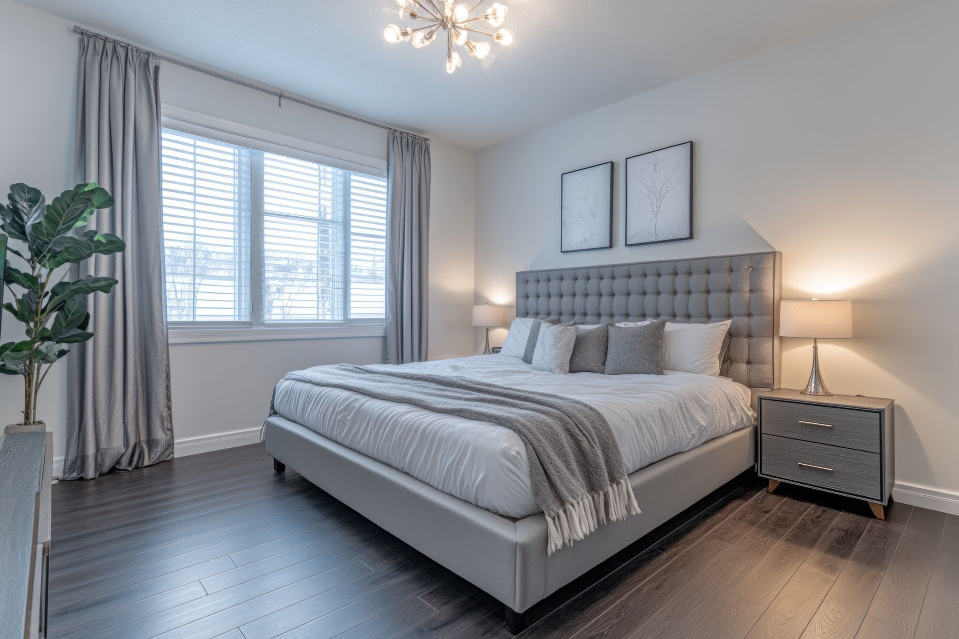 Bedroom with gray bed, nightstand, and curtains; plant, artwork, and window with blinds.