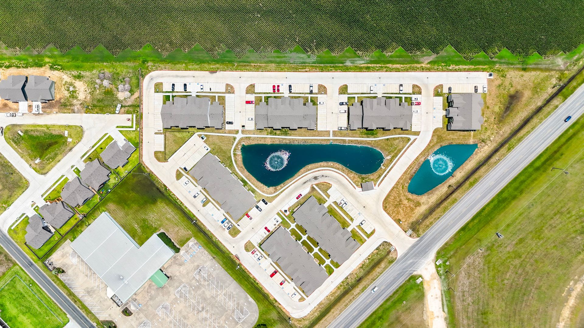 Aerial view of townhomes with a road and a field in the background. Green grass and a small pond are in the foreground.