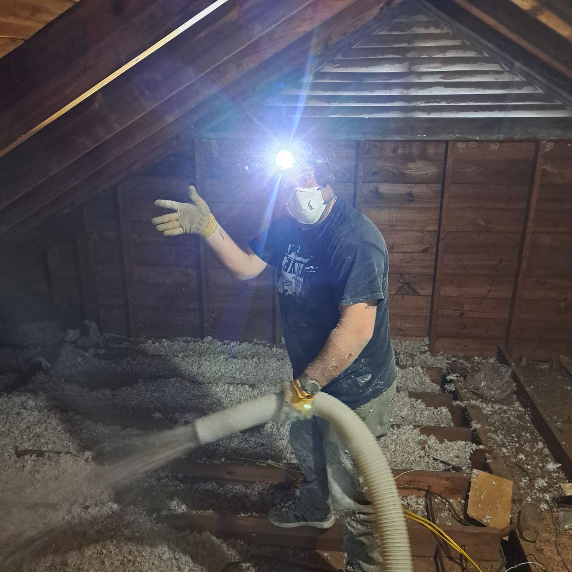 Worker vacuuming attic insulation under a bright headlamp, with dust hose and exposed wooden rafters.