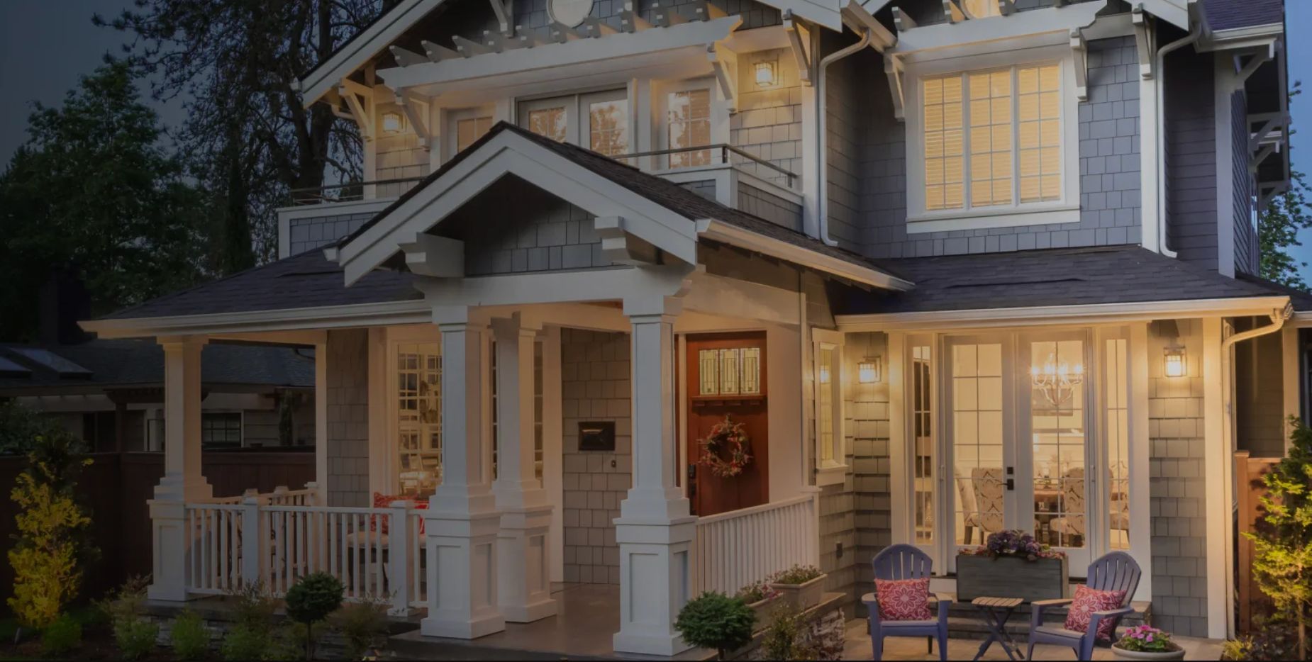White two-story house with a covered porch, lit windows, and outdoor chairs at dusk