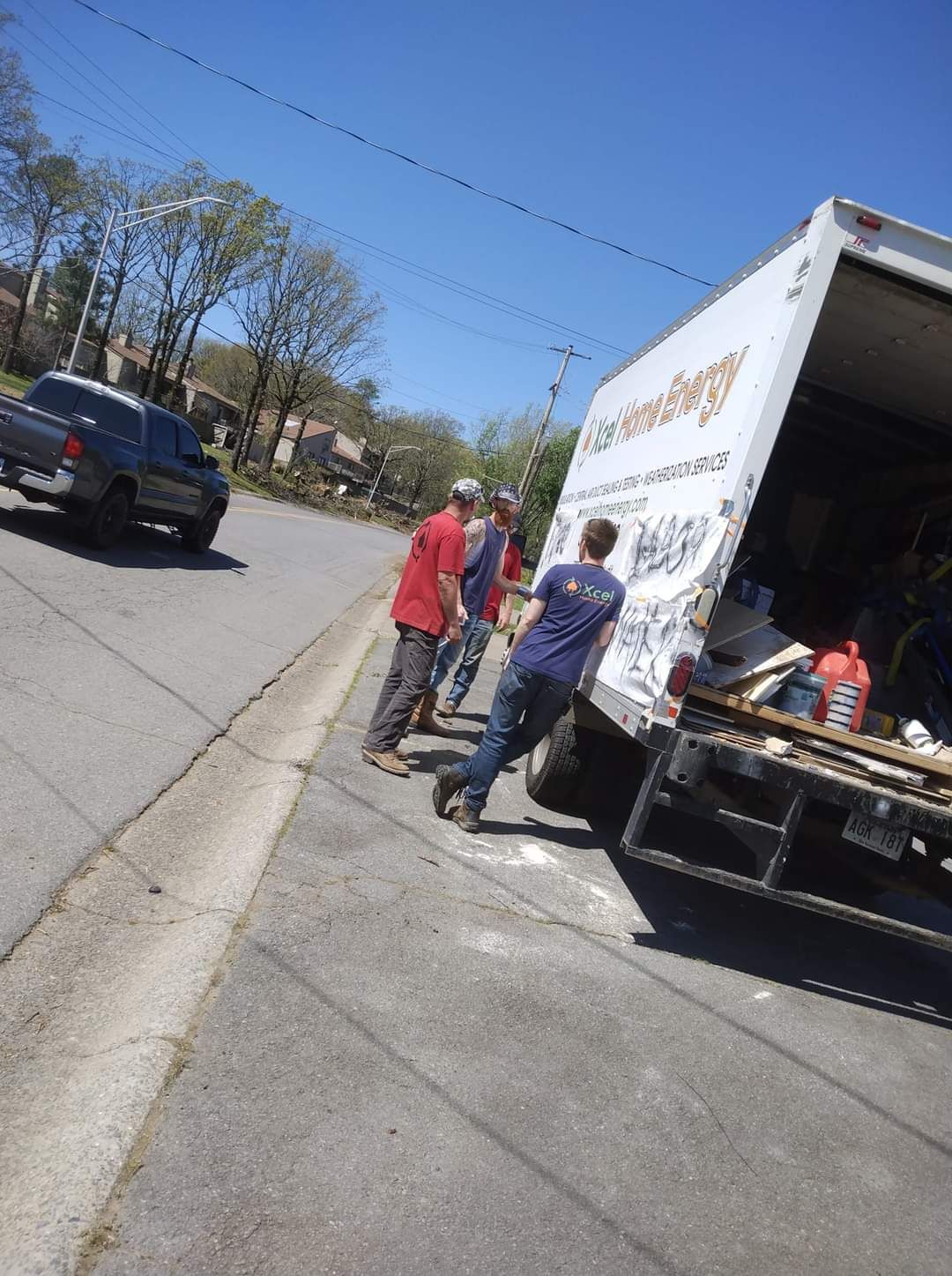 People unloading a truck on a sunny roadside beside parked cars and trees
