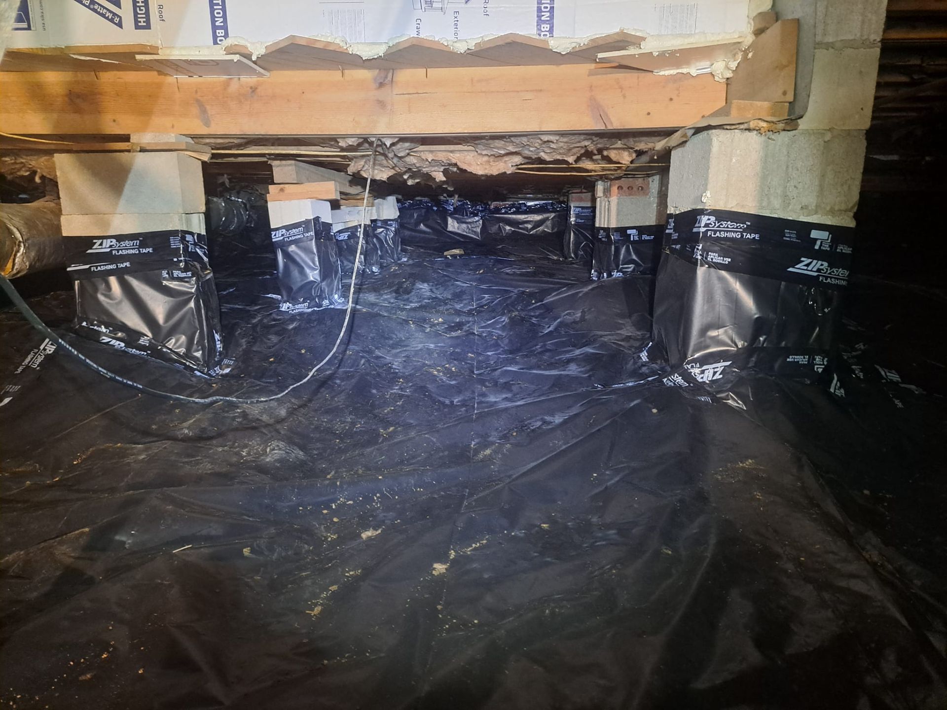 Basement crawlspace covered in black plastic sheeting beneath exposed floor joists and insulation.