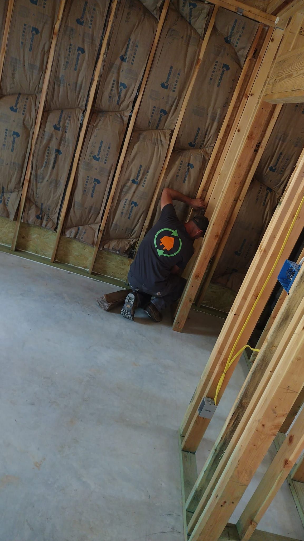 Worker crouching inside an unfinished room with exposed wooden framing and a concrete floor.